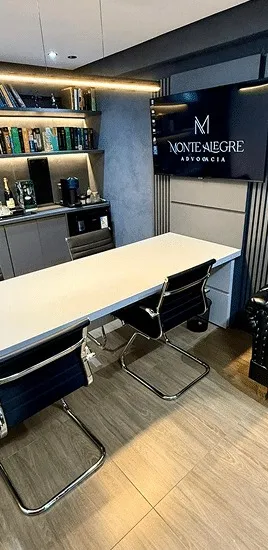Sala de reunião do escritório Monte Alegre, com mesa branca, cadeiras de couro preto, estante de livros e o logotipo na tela
